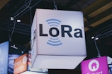 A brightly lit cube with the text 'LoRa' hangs from a ceiling, surrounded by various other illuminated signage. Industrial framework and lighting are visible in the background, suggesting a trade show or exhibition setting.
