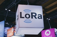 A brightly lit cube with the text 'LoRa' hangs from a ceiling, surrounded by various other illuminated signage. Industrial framework and lighting are visible in the background, suggesting a trade show or exhibition setting.