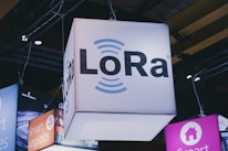 A brightly lit cube with the text 'LoRa' hangs from a ceiling, surrounded by various other illuminated signage. Industrial framework and lighting are visible in the background, suggesting a trade show or exhibition setting.