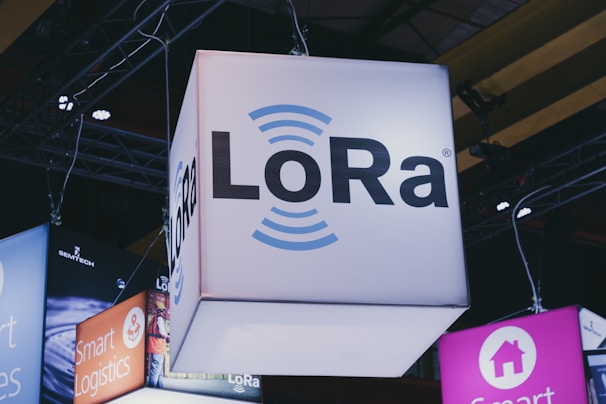 A brightly lit cube with the text 'LoRa' hangs from a ceiling, surrounded by various other illuminated signage. Industrial framework and lighting are visible in the background, suggesting a trade show or exhibition setting.