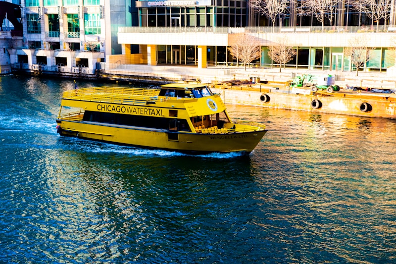 A bright yellow water taxi is cruising along a river, reflecting in the water below. The background features modern buildings with glass facades and a dock area with some trees and construction equipment. The river water contrasts with the vivid color of the water taxi.
