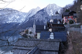 A picturesque alpine village with wooden and stone houses nestled beside a serene lake, surrounded by snow-capped mountains. The church steeple stands out among the traditional buildings, and bare tree branches frame the scene. The architecture is traditional and reflects a quaint, peaceful atmosphere.