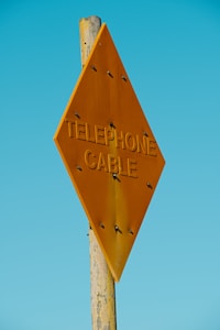 A wooden post supports a diamond-shaped, rusty orange metal sign with the words 'TELEPHONE CABLE' embossed on it. The background is a clear, bright blue sky, creating a vivid contrast with the sign.