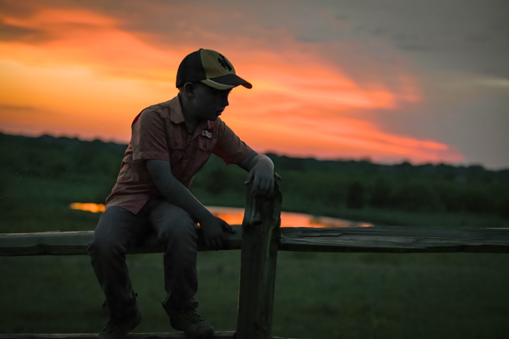 man in brown jacket and blue denim jeans sitting on brown wooden fence during sunset