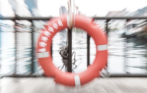 A motion-blurred image featuring a life preserver with text, situated on a metal railing by the water. The background includes buildings and urban structures reflected in the water, suggesting a waterfront or harbor setting.