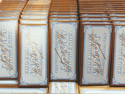 A collection of chocolate bars arranged neatly in rows, with vintage-style packaging featuring intricate blue designs and cursive script on a white background. The bars are labeled 'Vollmilch Extra Fein' indicating fine milk chocolate.