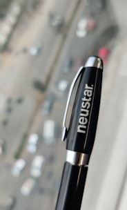 A black pen with a silver clip and the word 'neustar' written in white is held in focus against a blurred urban background, showing a road with several cars and buildings.