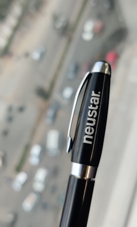 A black pen with a silver clip and the word 'neustar' written in white is held in focus against a blurred urban background, showing a road with several cars and buildings.
