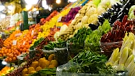 A vibrant and colorful market display showcasing a variety of fresh fruits and vegetables, including red, green, and yellow peppers, eggplants, oranges, and cabbages. The produce is stacked neatly in baskets and crates, creating an appealing and inviting scene.