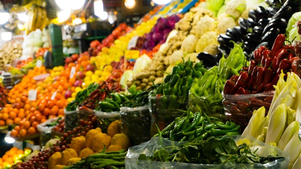 A vibrant and colorful market display showcasing a variety of fresh fruits and vegetables, including red, green, and yellow peppers, eggplants, oranges, and cabbages. The produce is stacked neatly in baskets and crates, creating an appealing and inviting scene.