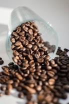A glass cup is tipped over with roasted coffee beans spilling out onto a surface. The beans are glossy and rich brown, catching light and creating a warm, inviting feel.