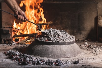 A hand is using a metal tool to tend to a fire in a rustic indoor setting. There is a large pile of ash and embers covering what appears to be a domed metal object on a stone surface. In the background, a bright fire is burning with logs and bricks visible.