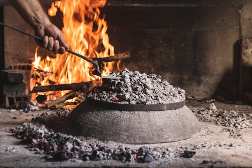 A hand is using a metal tool to tend to a fire in a rustic indoor setting. There is a large pile of ash and embers covering what appears to be a domed metal object on a stone surface. In the background, a bright fire is burning with logs and bricks visible.