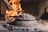 A hand is using a metal tool to tend to a fire in a rustic indoor setting. There is a large pile of ash and embers covering what appears to be a domed metal object on a stone surface. In the background, a bright fire is burning with logs and bricks visible.