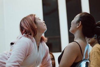 Two people are facing each other indoors, appearing to be in a playful or competitive interaction. One person has pink hair and is wearing a light-colored hoodie, while the other has dark hair tied back and is wearing a dark top. They seem to be leaning toward each other, possibly in a friendly challenge or game.