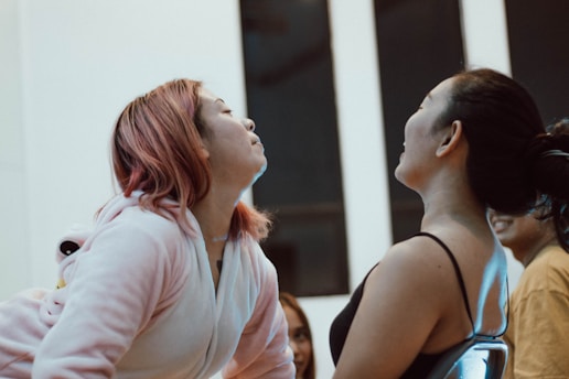 Two people are facing each other indoors, appearing to be in a playful or competitive interaction. One person has pink hair and is wearing a light-colored hoodie, while the other has dark hair tied back and is wearing a dark top. They seem to be leaning toward each other, possibly in a friendly challenge or game.