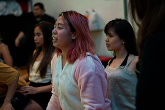 Several young people are gathered indoors, possibly engaged in a group activity or discussion. One person in the foreground, wearing a cozy, light-colored hoodie, has pink hair and appears to be speaking or singing. The mood seems relaxed with a casual setting.
