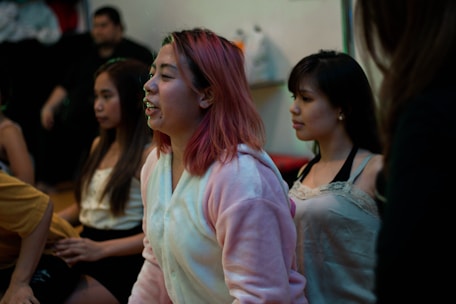 Several young people are gathered indoors, possibly engaged in a group activity or discussion. One person in the foreground, wearing a cozy, light-colored hoodie, has pink hair and appears to be speaking or singing. The mood seems relaxed with a casual setting.