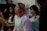 Several young people are gathered indoors, possibly engaged in a group activity or discussion. One person in the foreground, wearing a cozy, light-colored hoodie, has pink hair and appears to be speaking or singing. The mood seems relaxed with a casual setting.