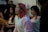 Several young people are gathered indoors, possibly engaged in a group activity or discussion. One person in the foreground, wearing a cozy, light-colored hoodie, has pink hair and appears to be speaking or singing. The mood seems relaxed with a casual setting.