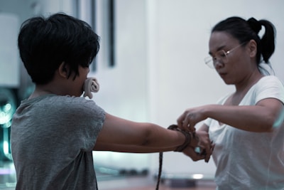 Two individuals are engaged in an activity where one person is tying the wrists of the other with a rope. Both are focused, and the setting appears to be indoors with soft lighting.