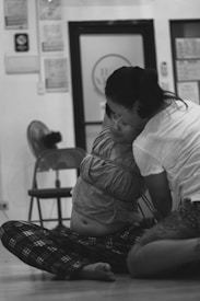 Two people are engaged in a bondage practice, with one person sitting on the floor, their torso wrapped in rope. The background includes a chair, a fan, and a door with signage, suggesting an indoor setting.