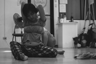 A woman sits on a wooden floor with her legs crossed, stretching her arms behind her head. She wears a loose top and plaid pants, appearing relaxed. In the background, there are chairs, a fan, and various objects on a counter, including bottles and cleaning supplies.