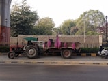 A tractor with a trailer is parked on a roadside. The trailer carries bags wrapped in pink material. Trees and foliage are visible in the background, along with a wall and a fence. A scooter is parked nearby.