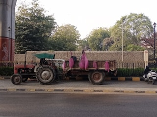 A tractor with a trailer is parked on a roadside. The trailer carries bags wrapped in pink material. Trees and foliage are visible in the background, along with a wall and a fence. A scooter is parked nearby.