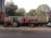 A tractor with a trailer is parked on a roadside. The trailer carries bags wrapped in pink material. Trees and foliage are visible in the background, along with a wall and a fence. A scooter is parked nearby.