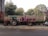 A tractor with a trailer is parked on a roadside. The trailer carries bags wrapped in pink material. Trees and foliage are visible in the background, along with a wall and a fence. A scooter is parked nearby.