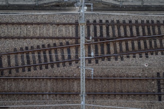 Aerial view of a railway track intersection. The tracks are laid on a bed of gravel, with several rails running parallel and others diverging at intersecting points. A metal structure appears to run vertically across the image, possibly part of an overhead power system.