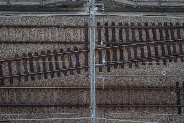 Aerial view of a railway track intersection. The tracks are laid on a bed of gravel, with several rails running parallel and others diverging at intersecting points. A metal structure appears to run vertically across the image, possibly part of an overhead power system.