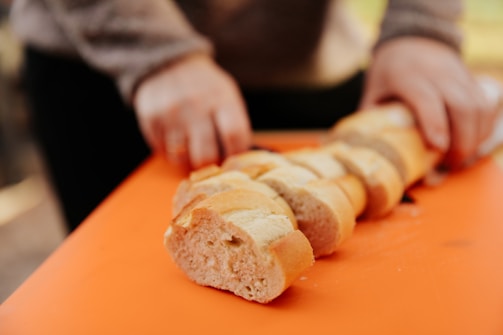 Two hands are slicing a baguette on an orange cutting board. The bread is cut into even slices, and the focus is on the process of slicing. The background is blurred, giving emphasis to the bread and hands.