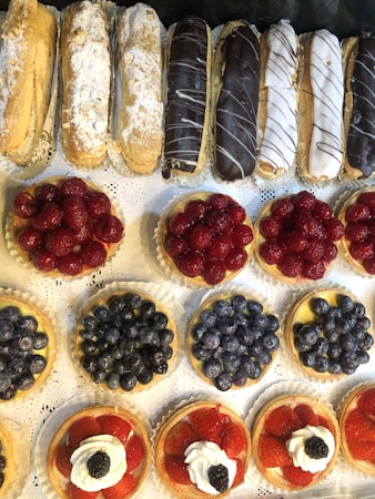 An assortment of delicious pastries is displayed on a white lace-patterned surface. The top row features eclairs with different toppings such as powdered sugar and chocolate glaze. Below, several fruit tarts are meticulously arranged, each topped with either raspberries, blueberries, or strawberries with a dollop of cream and a blackberry.
