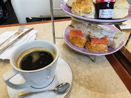 A tiered tray containing various food items including scones, sandwiches, croissants with salmon, and a jar of strawberry preserves. A cup of black coffee sits on the table beside a saucer with a teaspoon and a fork and knife placed on a napkin.