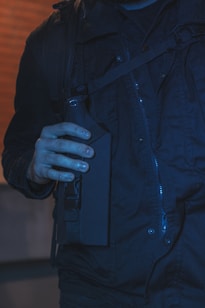 A person wearing a dark jacket holds a tactical-looking object, possibly a piece of equipment or gear, against a brick wall background.