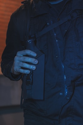 A person wearing a dark jacket holds a tactical-looking object, possibly a piece of equipment or gear, against a brick wall background.