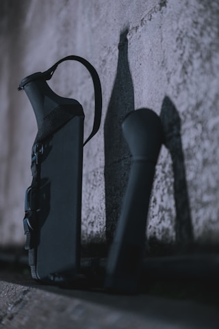 A dark and moody scene featuring two dark objects leaning against a textured concrete wall. The lighting casts strong shadows, emphasizing the rough surface of the wall and the shapes of the objects.