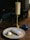 A candle on a brass holder sits on a wooden table beside a plate featuring a pastry dusted with powdered sugar and a scoop of ice cream. A small blue dish with a lid is also on the table. The dimly lit background suggests a cozy, intimate setting.