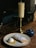 A candle on a brass holder sits on a wooden table beside a plate featuring a pastry dusted with powdered sugar and a scoop of ice cream. A small blue dish with a lid is also on the table. The dimly lit background suggests a cozy, intimate setting.