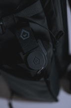 A close-up view of a black backpack featuring a rugged strap with a buckle. The strap has detailed stitching and a small logo imprinted on a section of fabric. The lighting creates a dark, moody atmosphere, highlighting the texture of the materials.