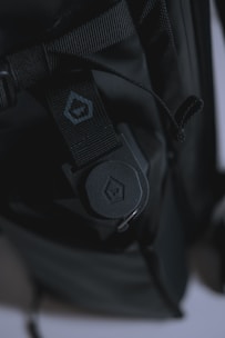 A close-up view of a black backpack featuring a rugged strap with a buckle. The strap has detailed stitching and a small logo imprinted on a section of fabric. The lighting creates a dark, moody atmosphere, highlighting the texture of the materials.