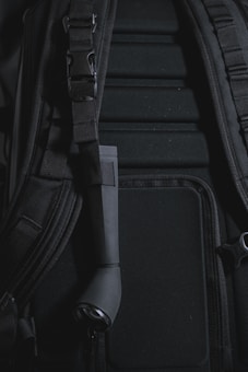 A close-up view of a backpack with visible padded sections and adjustable straps. There is a black cylindrical object attached to the strap, which appears to be a small flashlight.