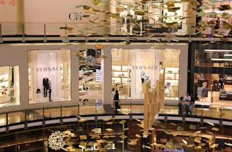 An interior view of a modern shopping mall showing multiple levels. Storefronts, including Versace, are visible with mannequins displaying luxury clothing. Golden decorations hang from the ceiling, creating a festive atmosphere. People can be seen walking and standing in the mall.