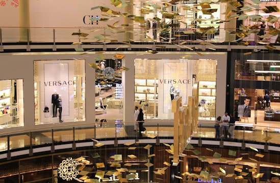 An interior view of a modern shopping mall showing multiple levels. Storefronts, including Versace, are visible with mannequins displaying luxury clothing. Golden decorations hang from the ceiling, creating a festive atmosphere. People can be seen walking and standing in the mall.