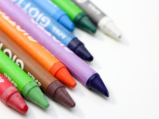 A close-up of an assortment of wax crayons in various colors, arranged diagonally on a white surface. The colors include red, green, brown, orange, purple, blue, and white. Each crayon has a slightly worn appearance and distinct labeling.