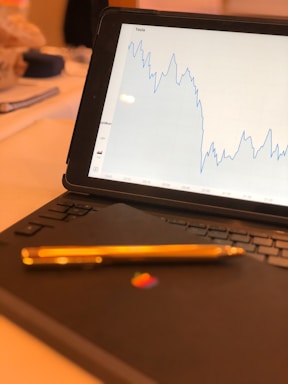 A tablet displaying a line graph with fluctuating data labeled as Tesla is positioned on a keyboard cover. In the foreground, a gold pen lies on top of the cover with a colorful logo visible.
