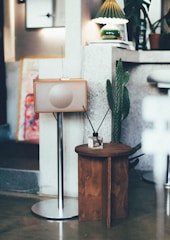 A retro-style radio stands on a sleek metal stand next to a small wooden table. A cactus plant adds a touch of greenery, positioned beside the table and supported by a stone wall. A green and white lampshade provides soft lighting, complementing the overall vintage and cozy atmosphere.