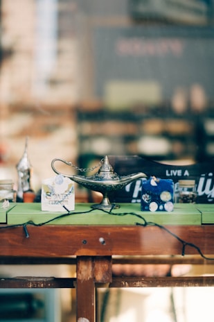 A vintage metal lamp with intricate designs prominently displayed on a wooden table, surrounded by various small items including a box labeled 'soap' and other decorative pieces. The background is blurred, suggesting an indoor or storefront setting.